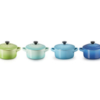 Set di quattro cocotte di diverse tonalità: verde lime, blu chiaro, blu scuro, con coperchi neri e manici laterali.
