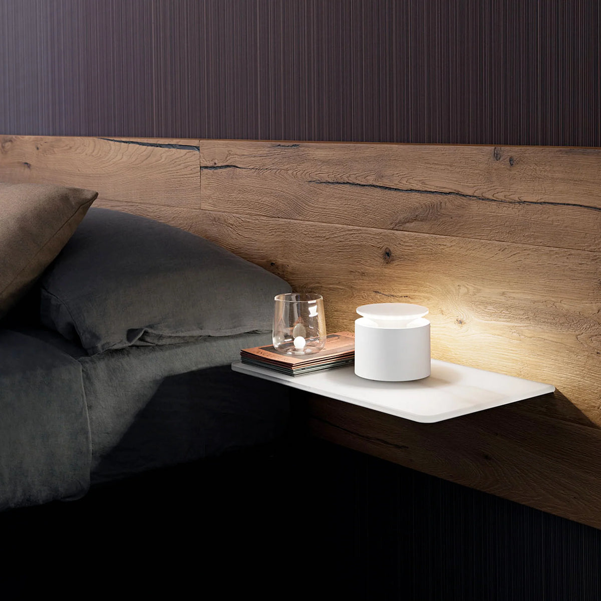 White cylindrical table lamp lit on white shelf with transparent glass and books, wood headboard and grey bed.
