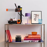 Metal shelf with orange and black 3D printer, purple table lamp, portrait painting, red book, light blue glass and lamp