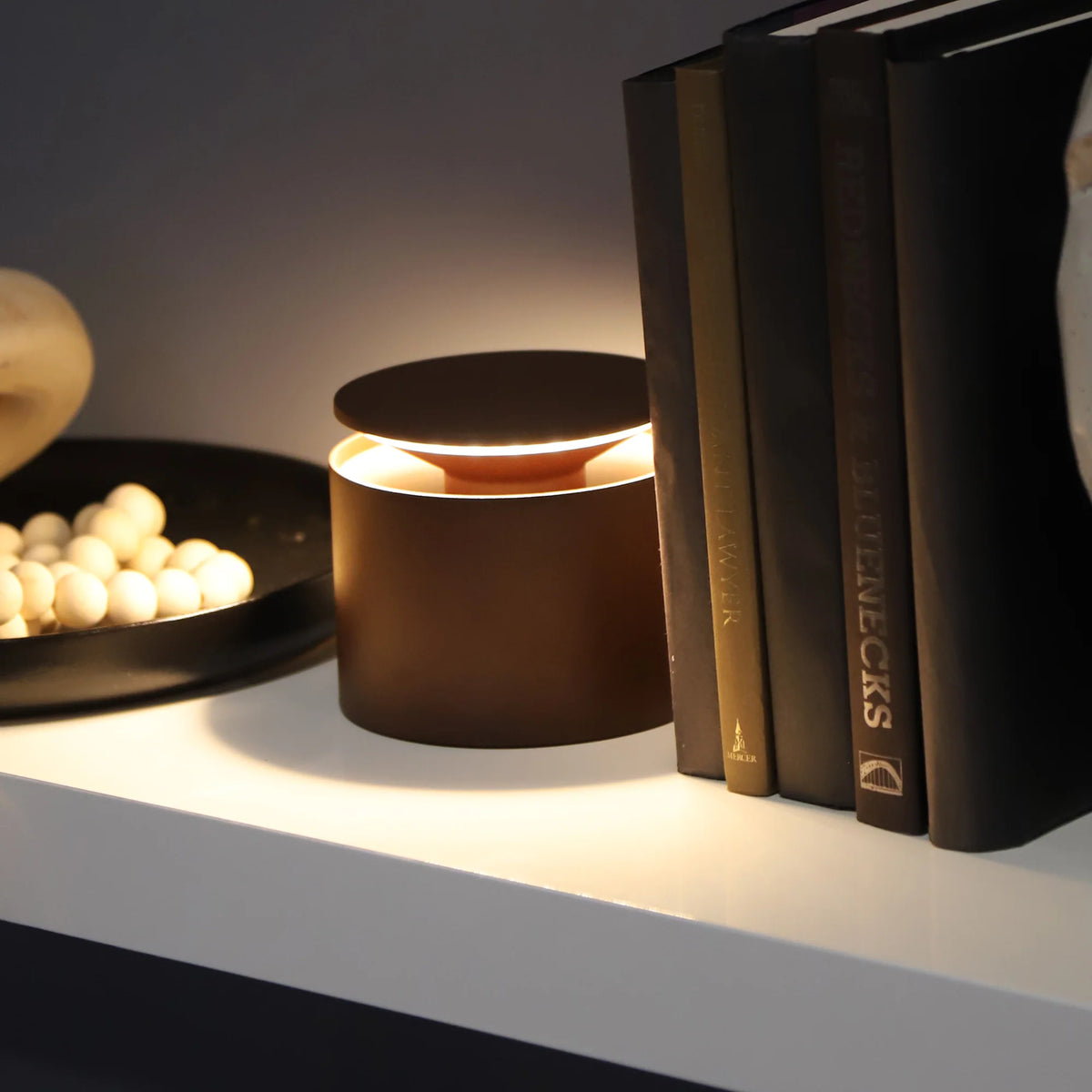Round brown table lamp with warm light, lit on white shelf next to books and a bowl with white spheres.