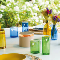 Cream cylindrical table lamp with warm light lit on white table among glasses and coloured glass bottles and dried flowers