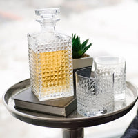 Carafe carrée en verre avec liquide doré, deux verres, livre gris et plante sur caisse métallique ronde