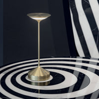 Lampada da tavolo in metallo dorato con base conica, stelo sottile e diffusore a forma di disco rovesciato su tavolo tondo a righe nere e