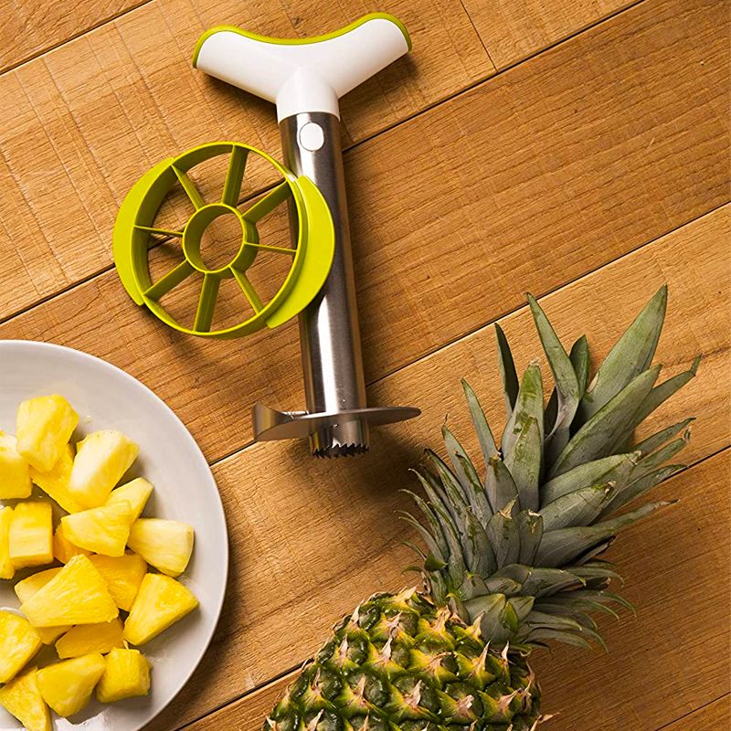 Cortador de piña de acero inoxidable con mango blanco y verde, junto a rodajas de piña en un plato y una piña entera.