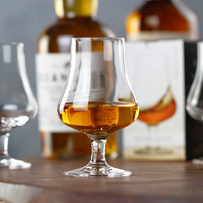 Verre transparent avec whisky ambré sur table en bois, deux bouteilles et un autre verre vide flou en arrière-plan