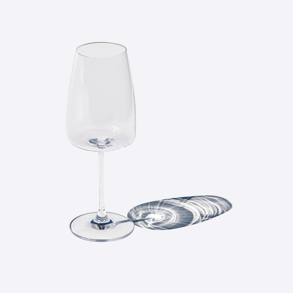 Verre transparent en verre, haut et élancé, avec base ronde et ombre projetée en spirale. Fond clair.