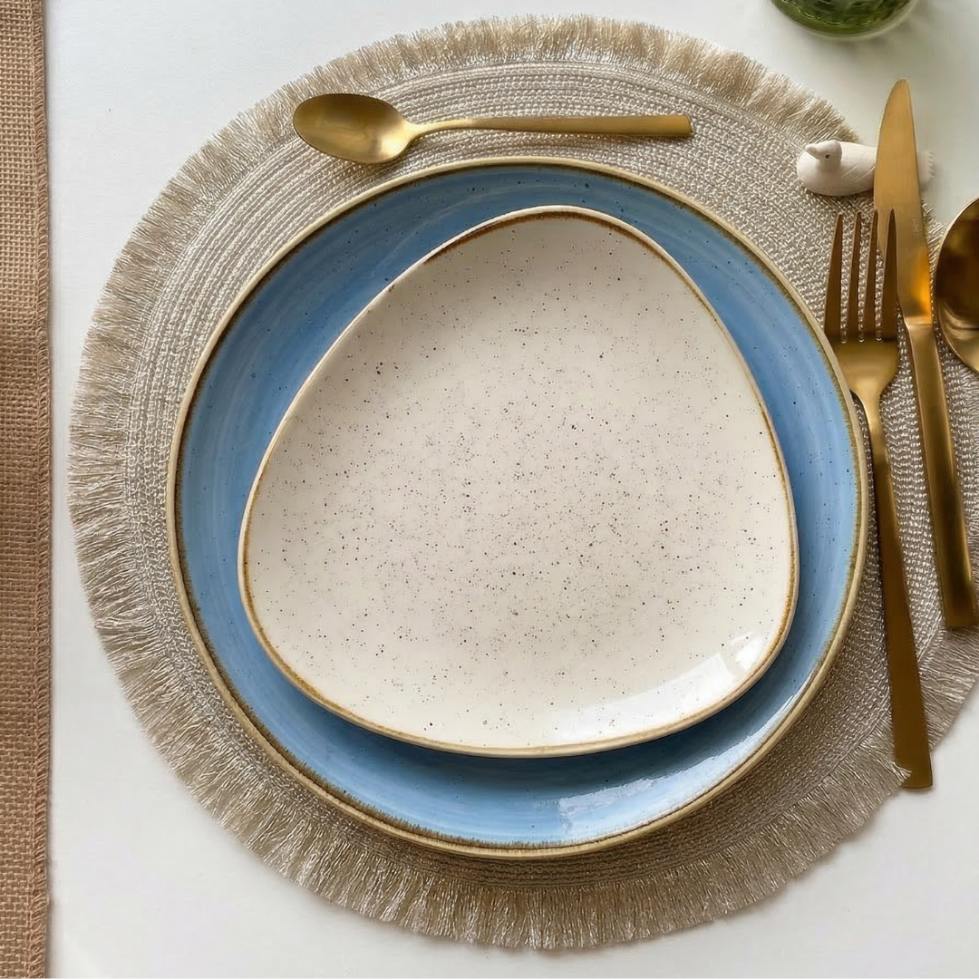 Two stacked plates on a beige charger with fringe, gold cutlery beside and a decorative object on top.