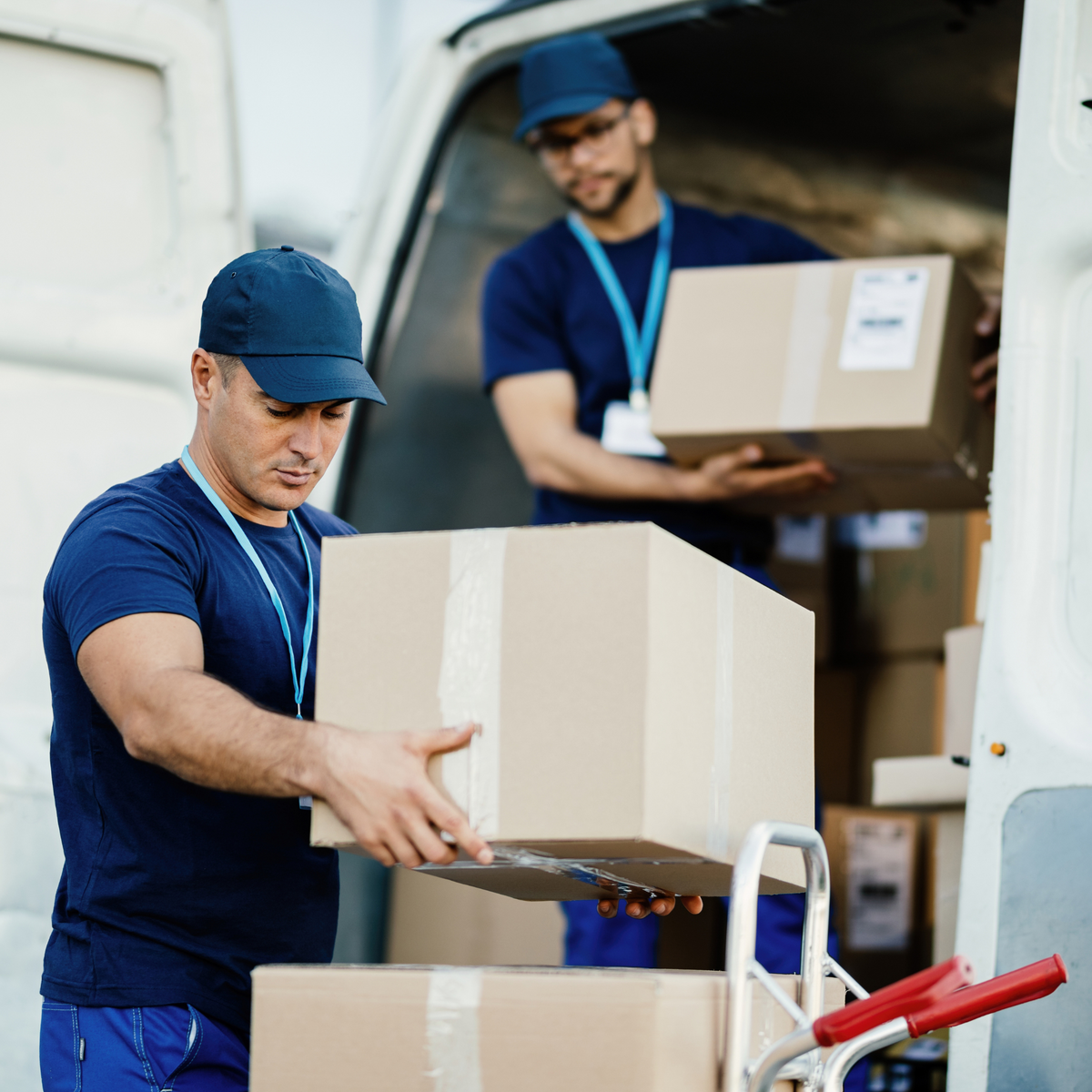 Two men in blue caps and blue uniforms loading and unloading cardboard boxes in a white van for