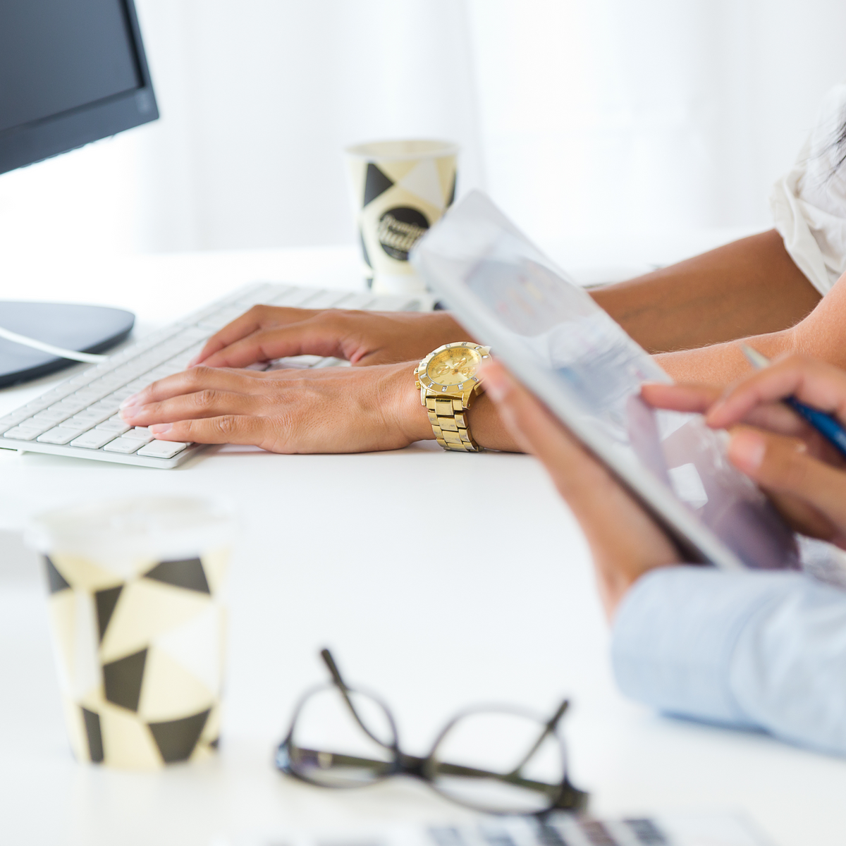 Two people's hands on white desk with keyboard, gold watch, glasses and black paper cups and