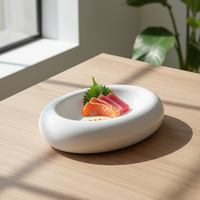 Assiette blanche brillante de forme ovale à bord épais contient tranches de sashimi rose et orange sur feuille verte, sur table en bois clair près de