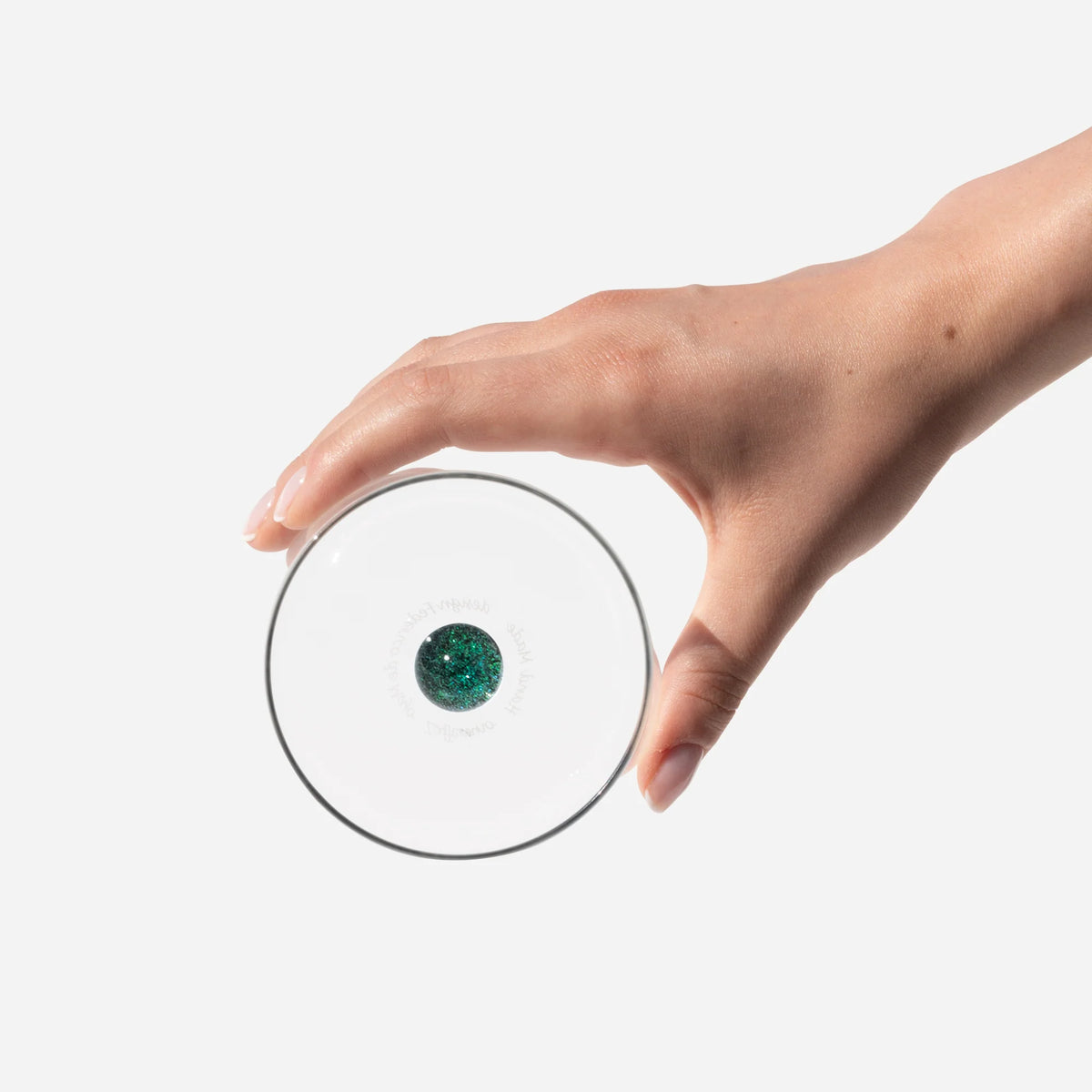 mano che tiene una lente circolare trasparente con un piccolo disco verde glitterato al centro su sfondo bianco uniforme