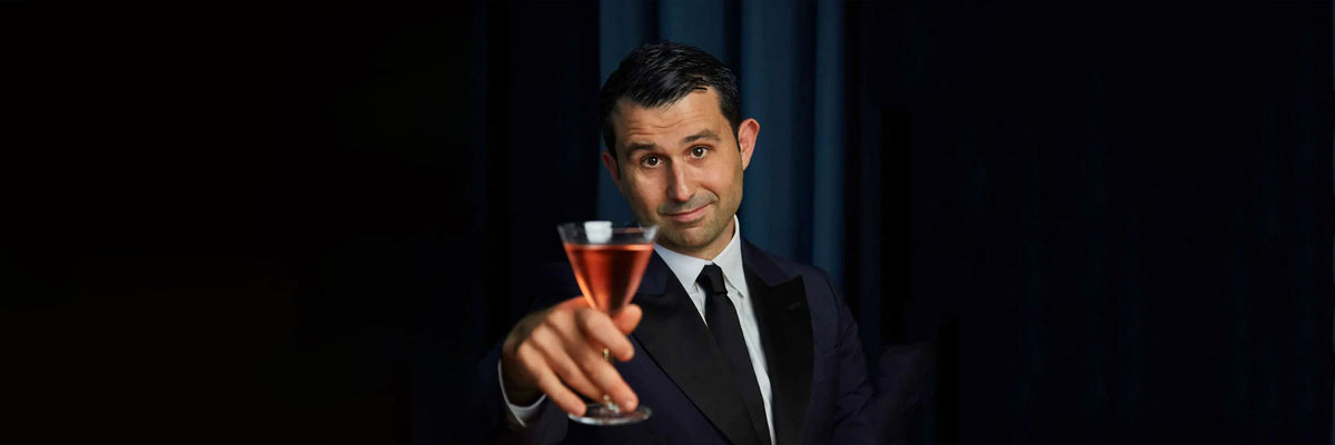 Uomo con abito scuro e cravatta che solleva un bicchiere da cocktail con liquido rosa e ghiaccio, sfondo nero.