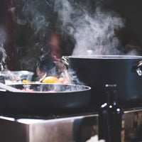 Zwarte pan en koekenpan met metalen handgrepen op kookplaat met stoom, wazige achtergrond met zwarte fles en ingrediënten