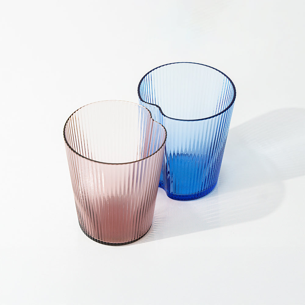 dos vasos de vidrio estriado, uno rosa claro y uno azul claro, con bordes irregulares, sobre superficie blanca con sombras ligeras