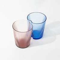 dos vasos de vidrio estriado, uno rosa claro y uno azul claro, con bordes irregulares, sobre superficie blanca con sombras ligeras