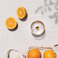 Two halved oranges, two whole oranges in beige cotton net and transparent water glass on light fabric