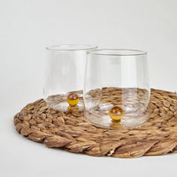 Two transparent glass tumblers with amber-colored ball at bottom on light brown woven coaster