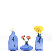 Trois vases en verre bleu transparent avec fleurs uniques blanches, orange et jaunes sur fond blanc minimaliste