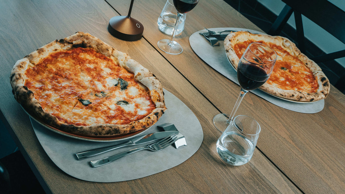 Two deep-dish margherita pizzas on light ceramic plates with raised rims, metal cutlery, red wine