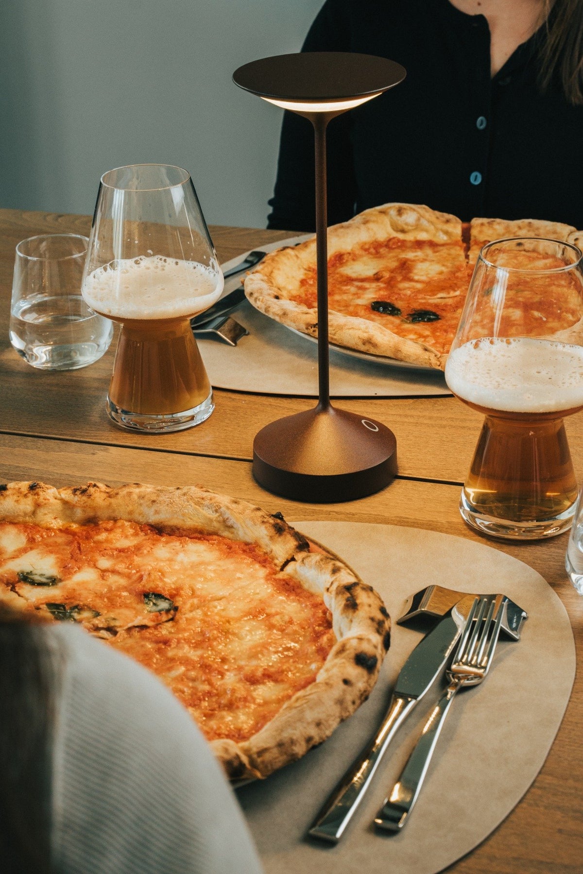 Pizza margherita su un tavolo di legno con bicchieri di birra e acqua, lampada moderna e posate accanto.