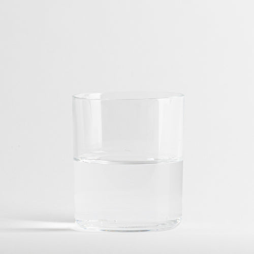 Bicchiere trasparente in vetro con acqua a metà, su sfondo bianco uniforme.