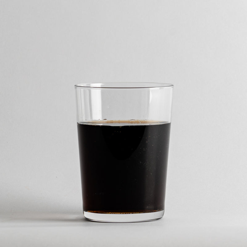 Vaso transparente de forma cilíndrica de cristal que contiene líquido oscuro sobre fondo blanco.