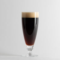Transparent tumbler with dark beer and beige foam on light grey background