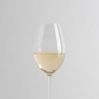 Transparante glazen wijnglas met lichte witte wijn en licht mousserend oppervlak op lichtgrijze achtergrond.
