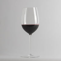Copa de vidrio transparente con pie con vino tinto a la mitad sobre fondo gris claro uniforme