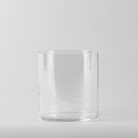 Vaso transparente de cristal liso y cilíndrico sin decoraciones sobre fondo gris claro.