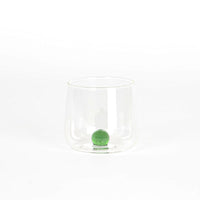 Bicchiere trasparente in vetro con base tonda e sfera verde all'interno, su sfondo bianco uniforme.