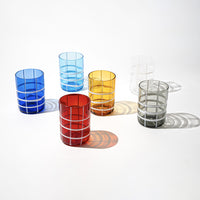 six verres en verre colorés en rouge, bleu, turquoise, jaune, gris et transparent avec motif à rayures blanches, sur fond blanc