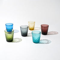 six verres en verre facettés de couleurs différentes sur fond blanc, vert, bleu clair, bleu, gris, jaune et rouge foncé, disposés en demi-cercle