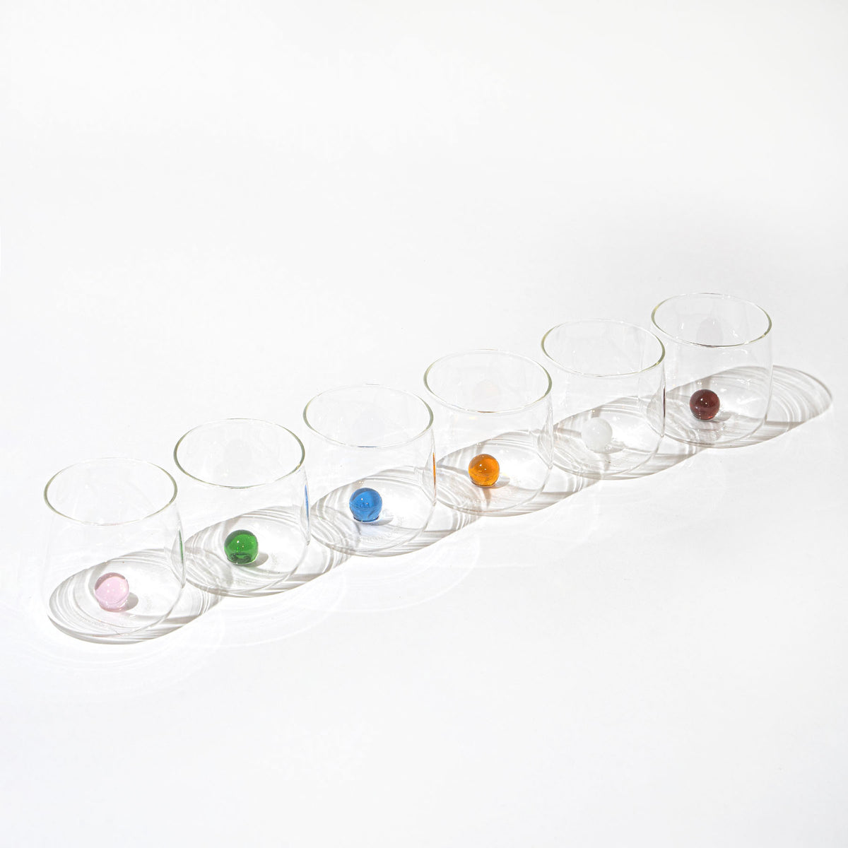 Set di sette bicchieri trasparenti in vetro, ognuno con una biglia colorata diversa all'interno, su sfondo bianco.