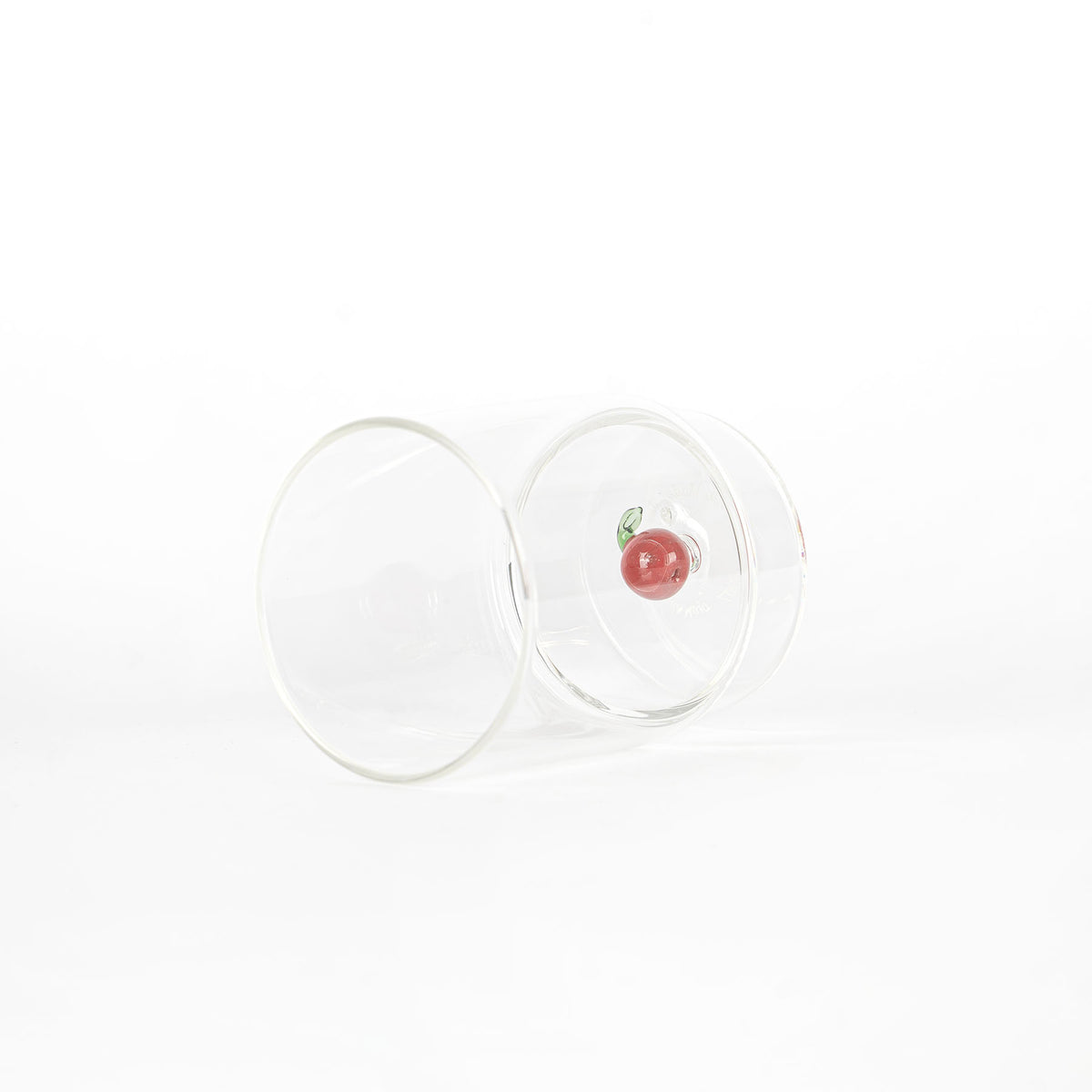 Bicchiere trasparente in vetro con decorazione a ciliegia rossa e foglia verde sul fondo, su sfondo bianco.