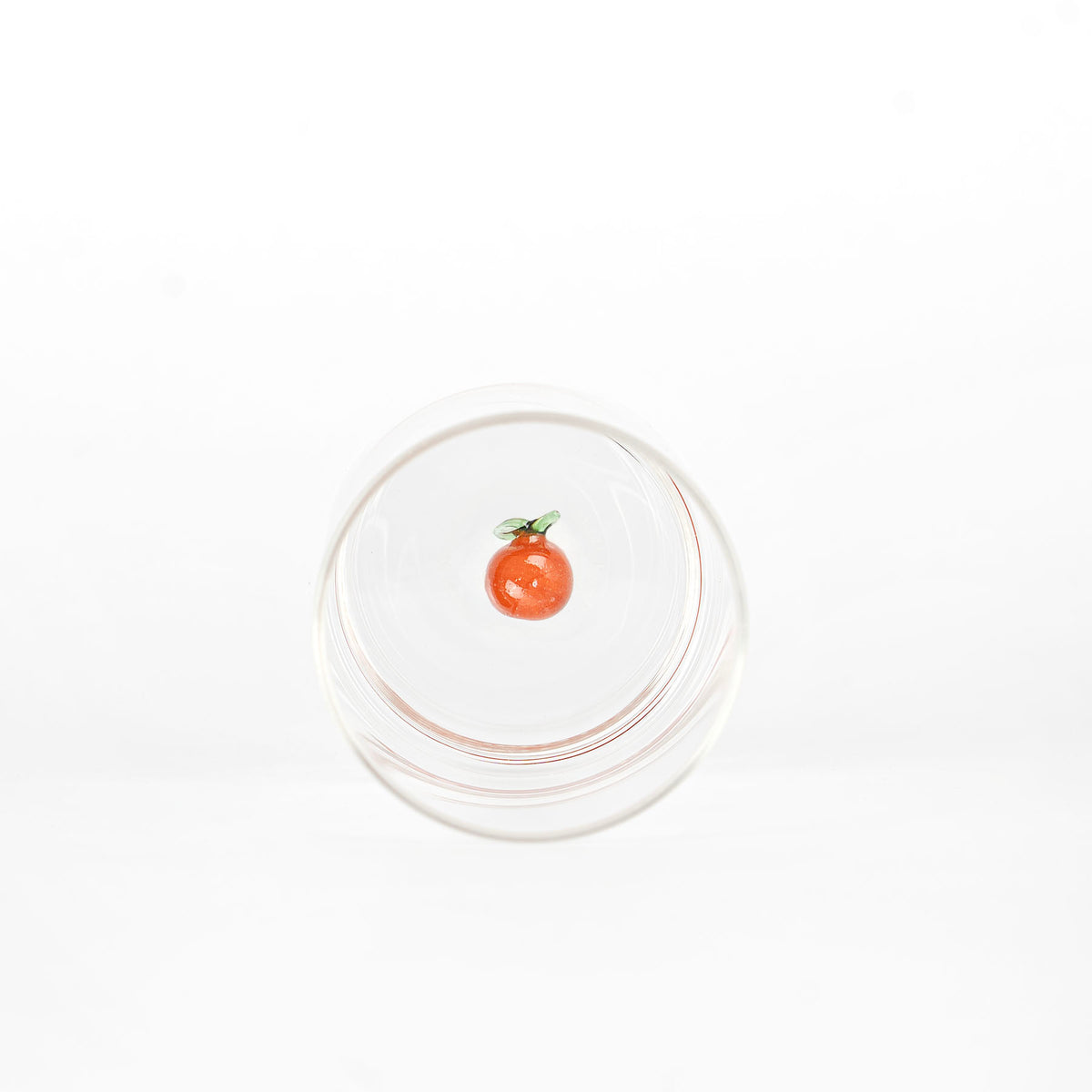 bicchiere di vetro trasparente rotondo con piccolo ornamento a forma di frutto arancione e foglie verdi sul fondo, sfondo bianco