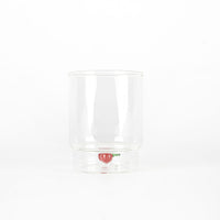 Bicchiere trasparente in vetro con base cava contenente decorazione a forma di ciliegia rossa con foglia verde, sfondo bianco.