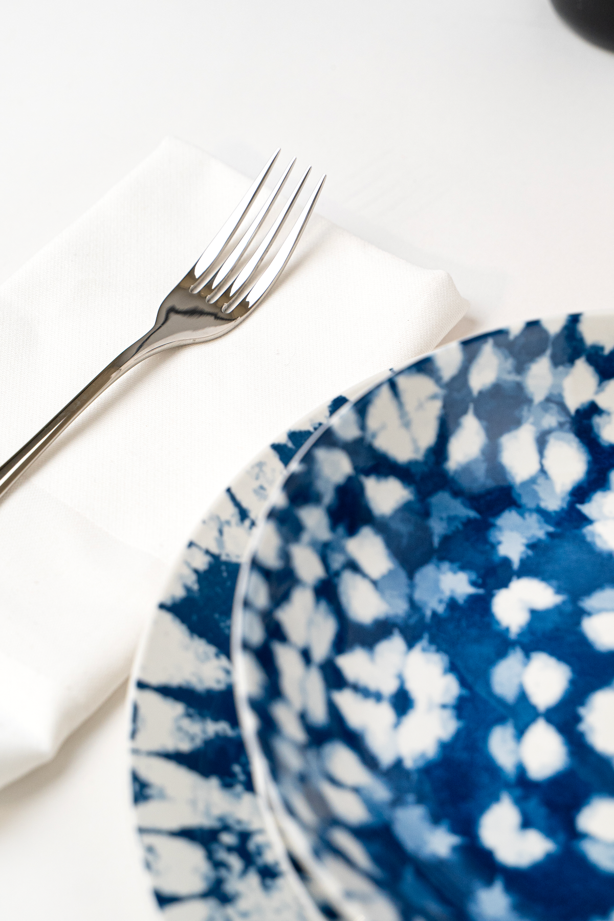 Blue and white decorated ceramic plate, next to glossy fork on white napkin. Light background.