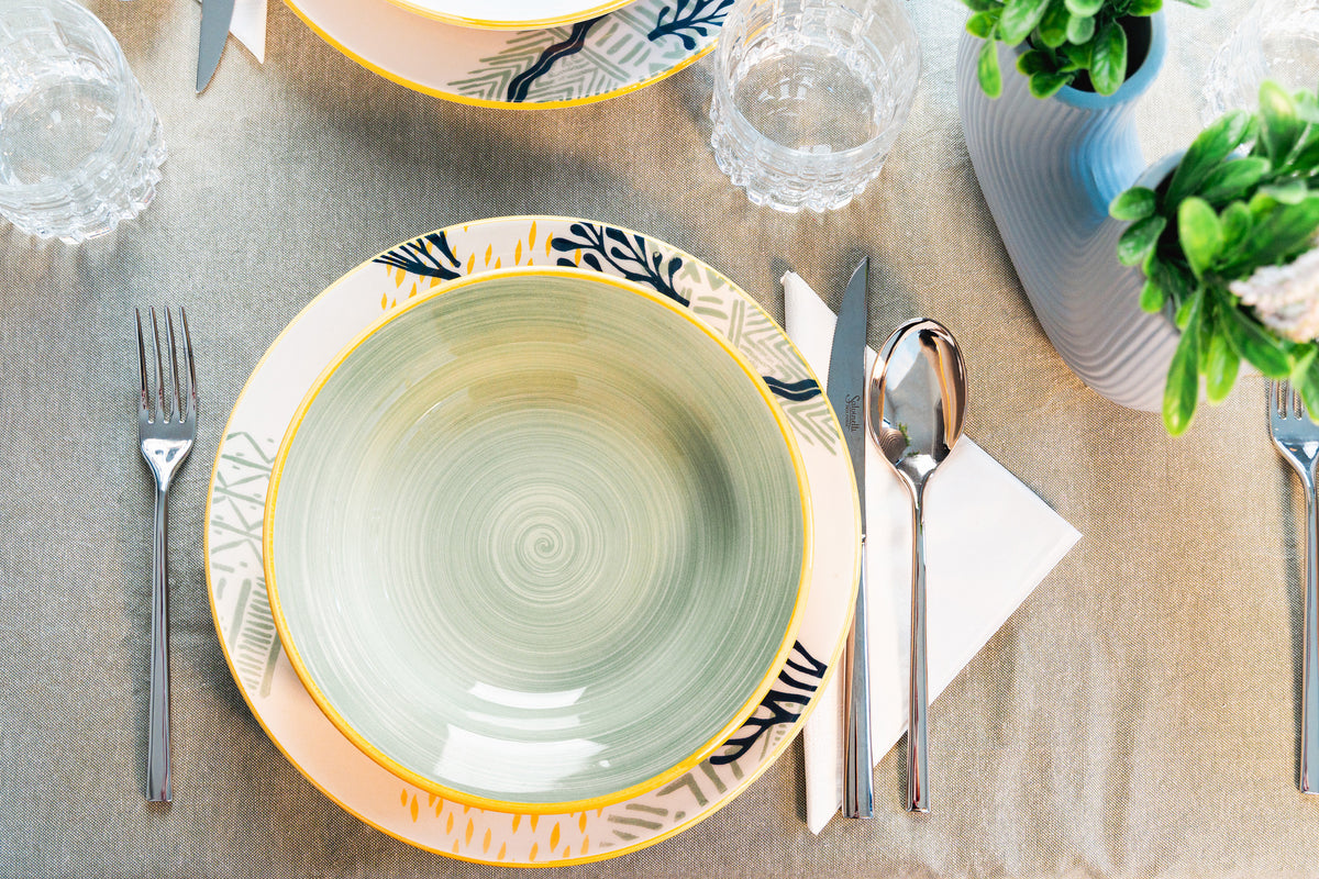Set di tavola elegante con piatti colorati, posate in metallo, bicchieri di vetro e un vaso decorativo con piante.