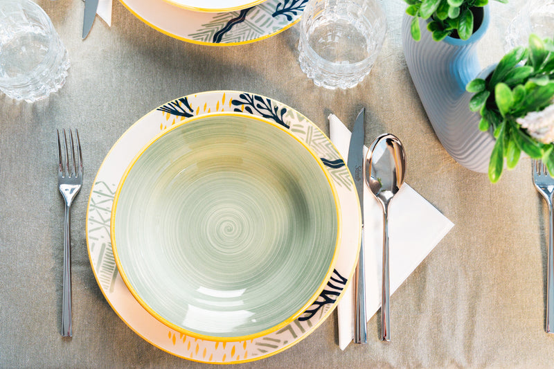 Set di tavola elegante con piatti colorati, posate in metallo, bicchieri di vetro e un vaso decorativo con piante.