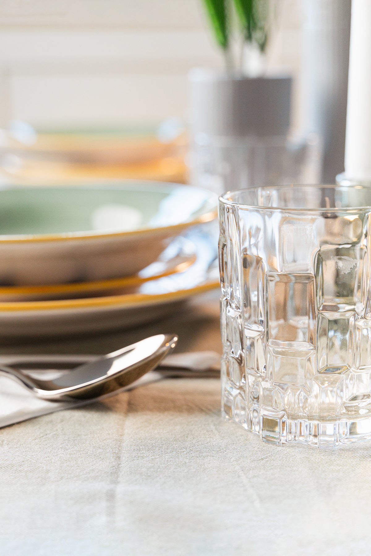 Dining table with green and white plates, transparent glass, metal cutlery and light tablecloth.