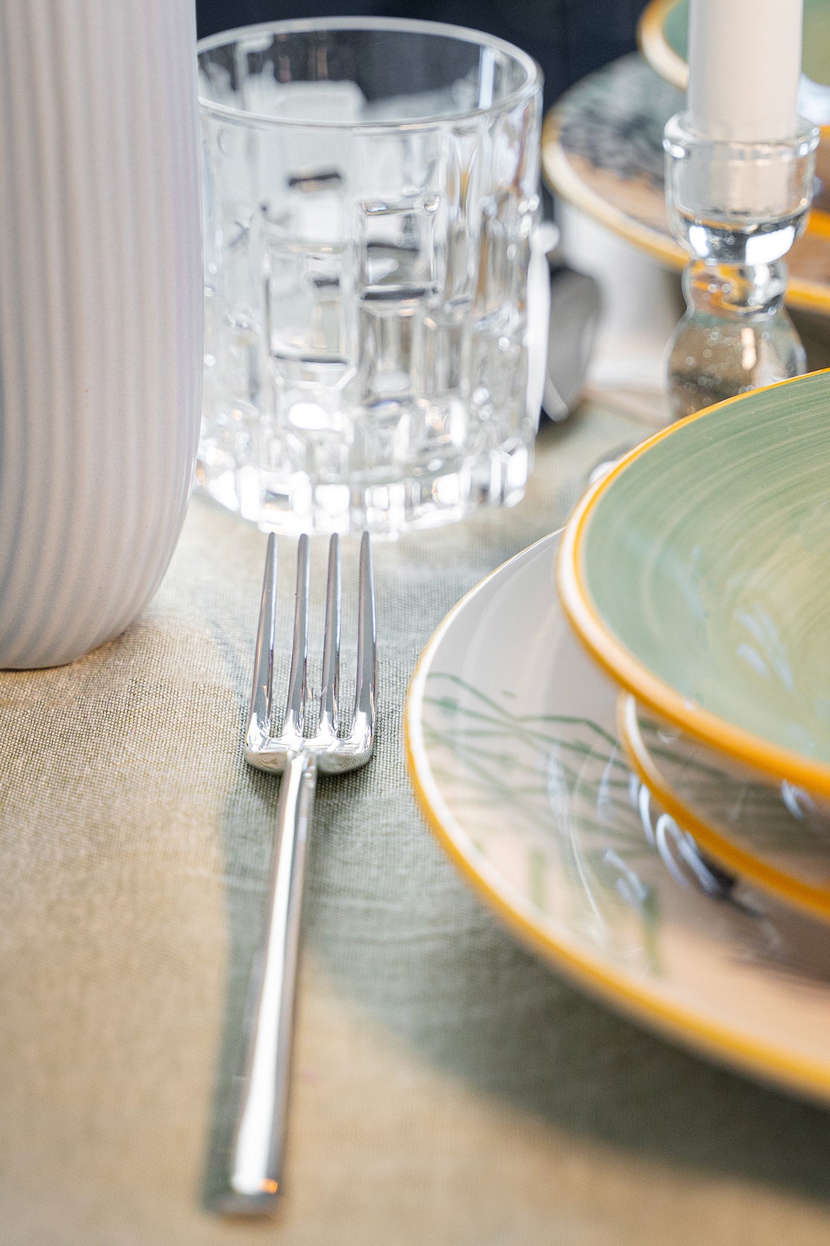 Fork and colorful plates on a beige tablecloth, a glass cup and a white vase are also visible. Soft lighting enhances the setting.