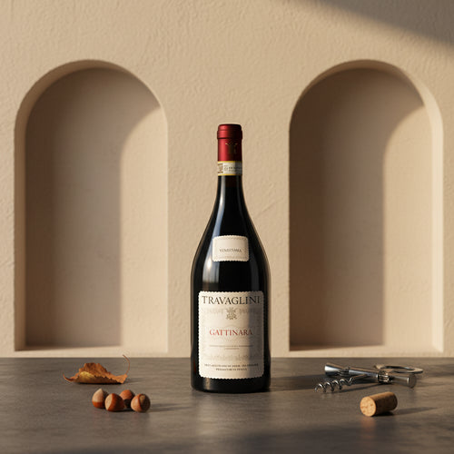 Bottiglia di vino rosso Travaglini Gattinara con etichetta bianca, posa su tavolo con noci, foglia secca, cavatappi e sughero, sfondo muro beige con