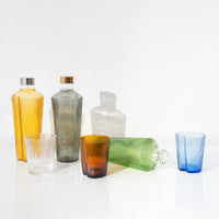 Botellas y copas de vidrio estriado transparente, naranja, verde, azul y gris sobre superficie blanca con fondo claro.