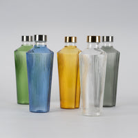 Fünf Glasflaschen mit geriffelter Oberfläche in Grün, Blau, Gelb, Transparent und Grau, mit goldenen und silbernen Metallverschlüssen auf neutralem Hintergrund.