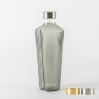 Graue transparente Glasflasche mit vertikal geriffelter Oberfläche und silbernem Metallverschluss, mit zwei extra gold- und silberfarbenen Verschlüssen auf Hintergrund