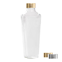 Transparente Glasflasche mit vertikalen Rillen, goldenem Verschluss, daneben zwei gold- und silberfarbene Verschlüsse.
