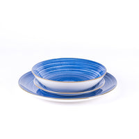 Ciotola bianca con bordo dorato e piatto blu con righe, entrambi in ceramica, posizionati uno sopra l'altro su sfondo bianco.