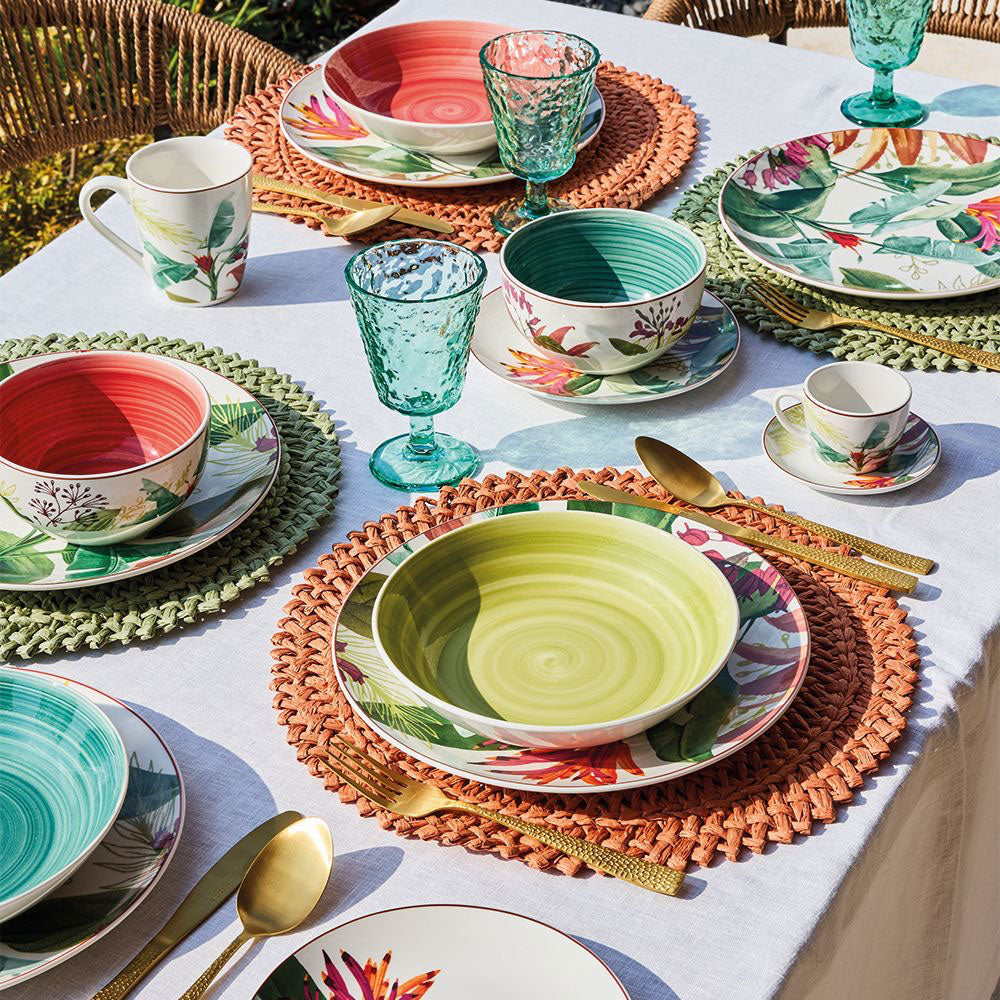 Tavola apparecchiata all'aperto con piatti, tazze e ciotole in ceramica colorata e bicchieri verdi in vetro, posate dorate.