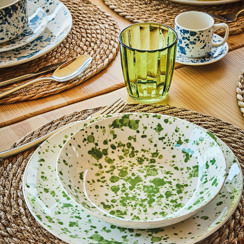 Set de platos de cerámica blanca con manchas verdes, vaso verde transparente, taza con platillo con manchas azules sobre mesa de madera con